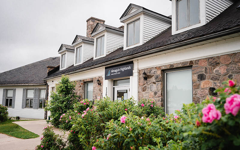 A view of the AHFHT's office in Burk's Falls, a stone building with 5 peaked windows in the roof, and pink flowers in front of the building.